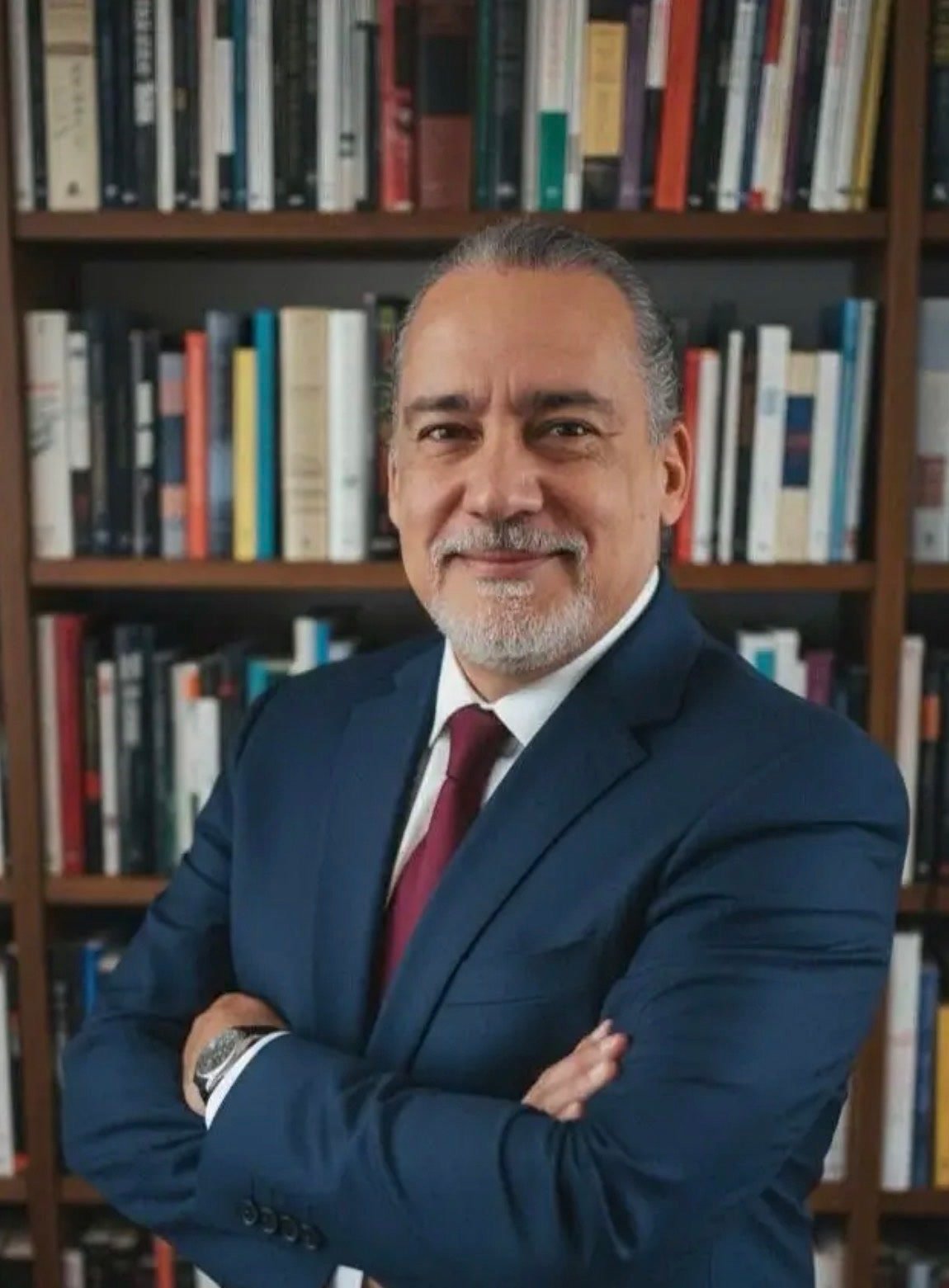 Dr. José Maurício, de terno azul e braços cruzados, diante de uma estante de livros